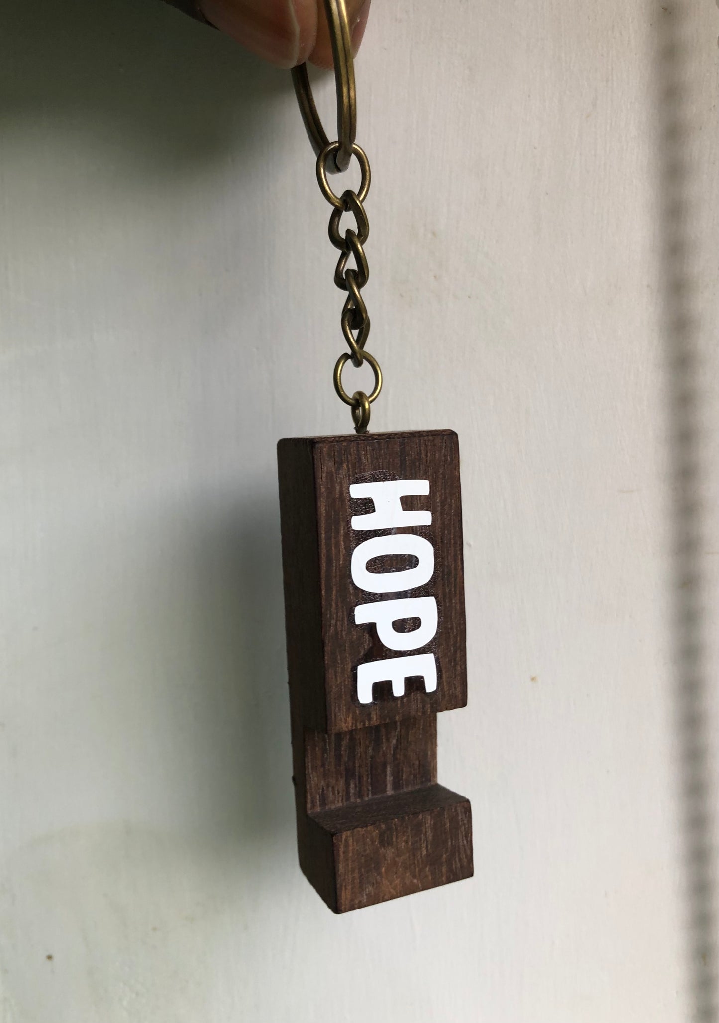 A dark brown wooden keychain charm spelling out the word 'HOPE' in white letters, attached to a metal key ring and chain, held against a plain off-white background.