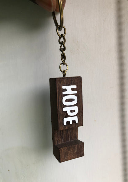 A dark brown wooden keychain charm spelling out the word 'HOPE' in white letters, attached to a metal key ring and chain, held against a plain off-white background.