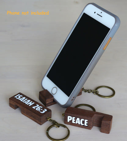 A silver smartphone propped upright on a small wooden block keychain holder engraved with 'ISAIAH 26:3'. Another matching wooden block keychain at the base reads 'PEACE'.