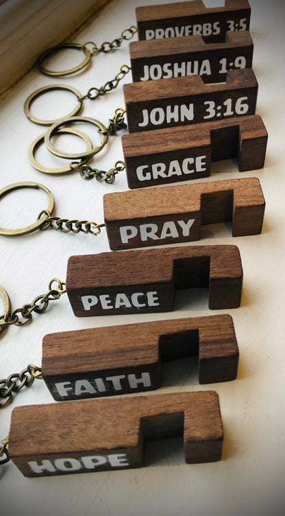 A set of wooden keychain puzzle tags with metal keys, each engraved with a different word of faith: PROVERBS 3:5, JOSHUA 1:9, JOHN 3:16, GRACE, PRAY, PEACE, FAITH, and HOPE, arranged diagonally on a light surface.
