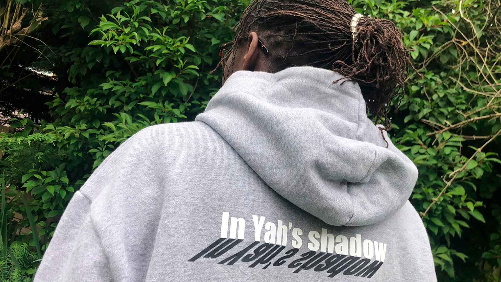 Person wearing a gray hoodie with text "In Yah's shadow" printed in white with a black shadow of the text on the back, standing in front of green foliage.